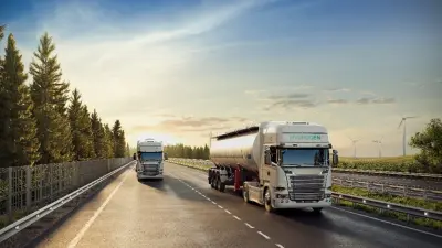 Two modern trucks drive on a sunny highway, surrounded by trees and wind turbines.